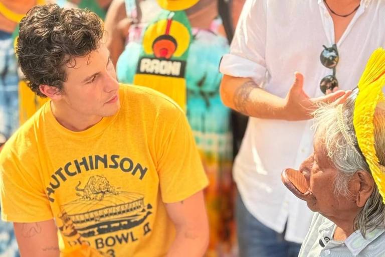 Jovem de camiseta amarela senta-se e segura a mão de idoso com faixa amarela na cabeça. Pessoas ao fundo observam a interação em ambiente iluminado.