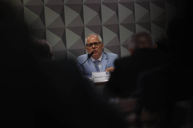 Homem de terno azul claro e óculos fala ao microfone em audiência pública. Fundo com painel geométrico cinza, público desfocado em primeiro plano.