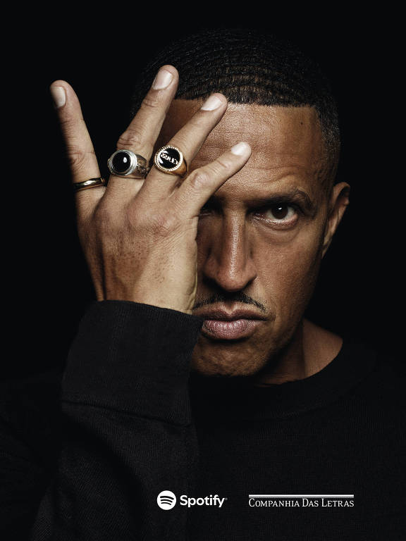 Homem com expressão séria, cabelo curto e ondulado, veste camiseta preta. Ele levanta a mão esquerda cobrindo o olho direito, usando três anéis grandes, um deles com símbolo de sorriso. Fundo preto.