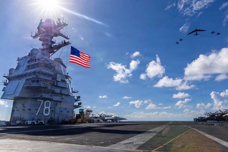 Porta-aviões USS Gerald R. Ford com número 78 visível na superestrutura, bandeira dos EUA hasteada, sob céu azul com nuvens. Esquadrilha aérea de cinco aviões voa em formação acima do convés.