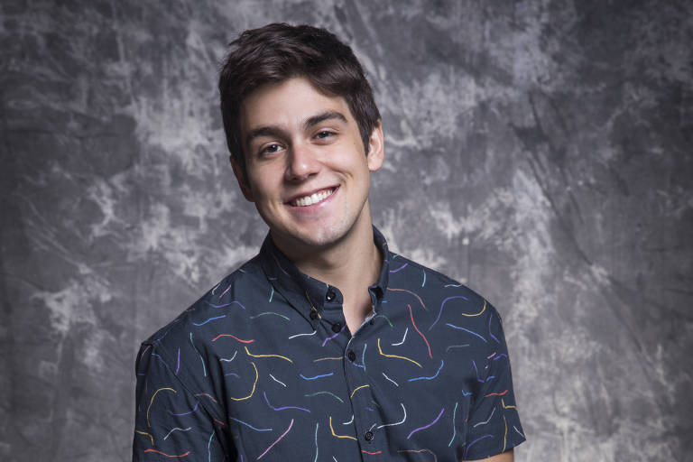 Homem branco posa sorrindo, vestido com camisa societal  de mangas curtas e calça vermelha 