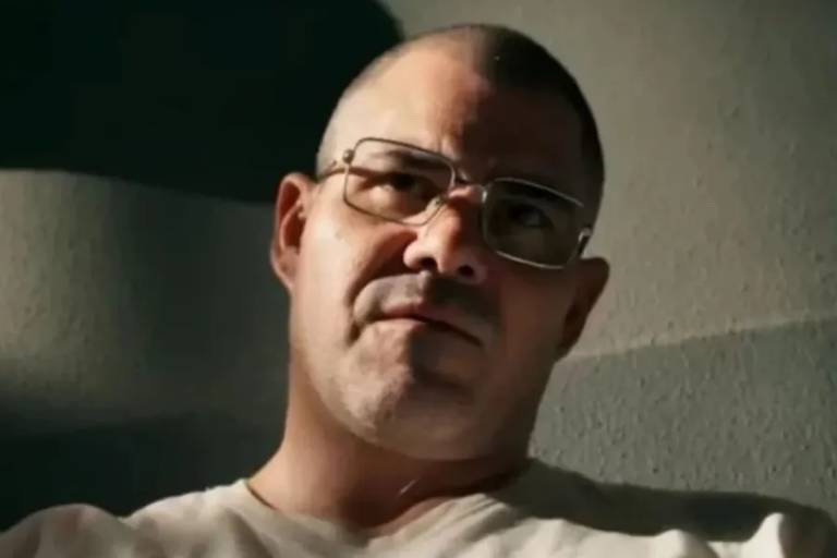 Homem de pele clara, cabeça raspada e óculos de armação metálica, olhando para frente com expressão séria. Fundo neutro com iluminação que destaca seu rosto.