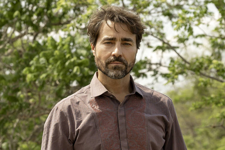 Homem de barba e cabelo curto está em pé sobre solo rochoso em área agrarian  com árvores ao fundo. Ele veste camisa marrom, calça jeans azul e botas cinza.