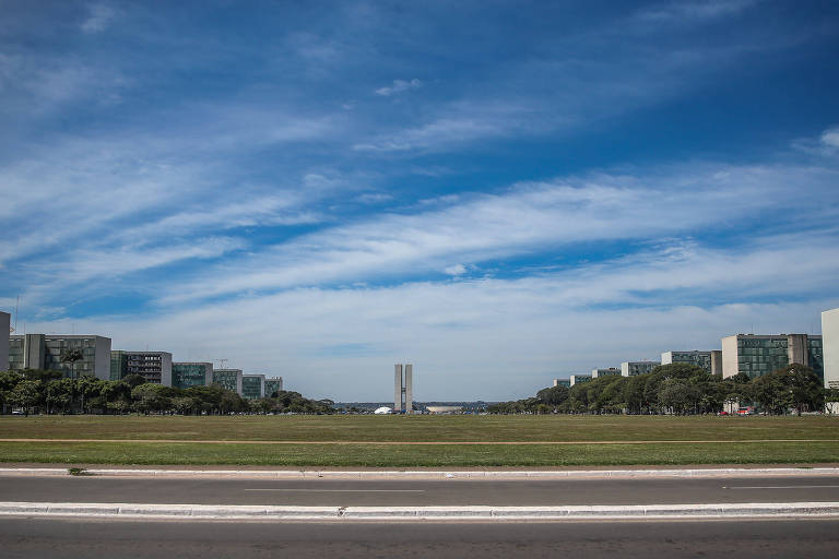 Eixo Monumental de Brasília com gramado amplo ao centro, ladeado por prédios modernos e árvores. Ao fundo, o Congresso Nacional com suas duas torres e cúpulas distintas sob céu azul com nuvens dispersas.