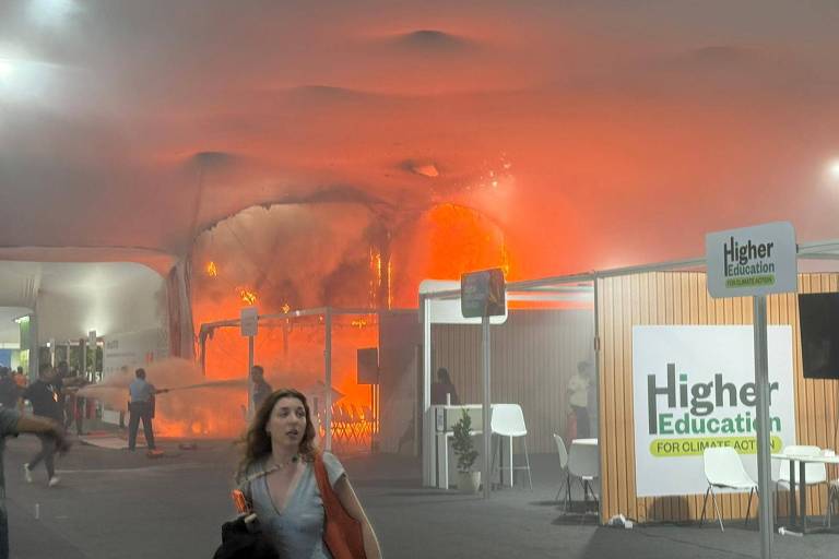 Fumaça densa e chamas alaranjadas saem de um estande em chamas durante evento de educação. Pessoas evacuam o local enquanto o fogo consome a estrutura.