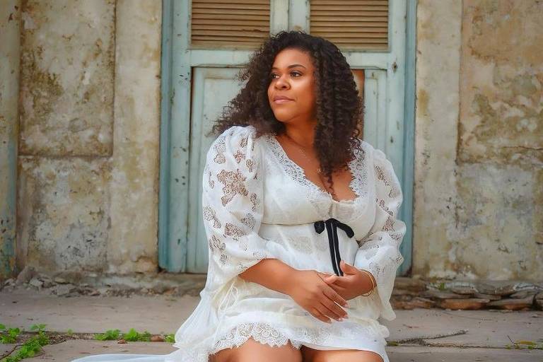 Mulher de cabelos cacheados sentada descalça em degraus de pedra, vestindo vestido branco com mangas bufantes e detalhes em renda, com fita preta na cintura. Ao fundo, porta azul clara com venezianas marrons em parede de textura envelhecida.