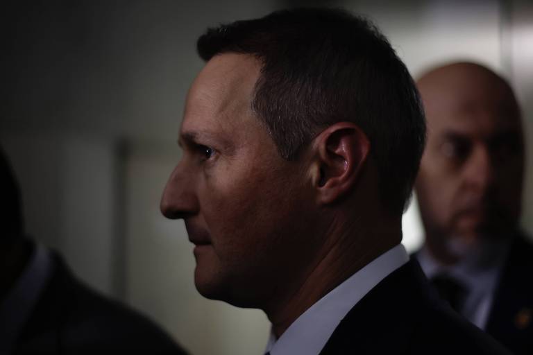 Homem de perfil com cabelo curto e terno escuro em ambiente interno pouco iluminado. Ao fundo, duas pessoas desfocadas, uma delas careca, também vestindo roupas formais.