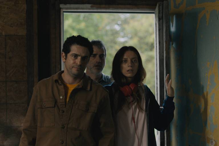 Three adults are near an open door indoors. A man on the left wears a brown shirt, in the center another man with a beard and gray hair, and a woman on the right with dark hair and a red scarf. The wall on the right has blue paint with yellow markings.