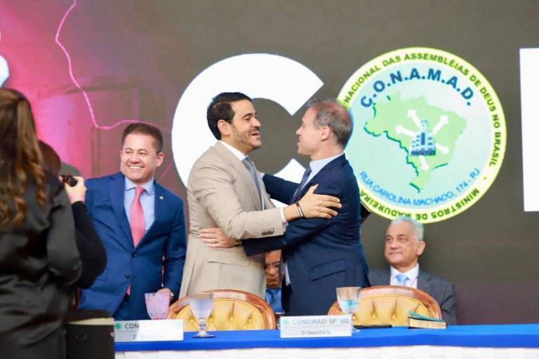 Dois homens de terno se abraçam no centro de uma mesa de conferência. Ao fundo, há um logotipo circular da CONAMAD com mapa do Brasil. À esquerda, um homem sorri e observa a cena. À direita, um homem mais velho está sentado, olhando para frente. Uma mulher com câmera fotografa a cena de costas.