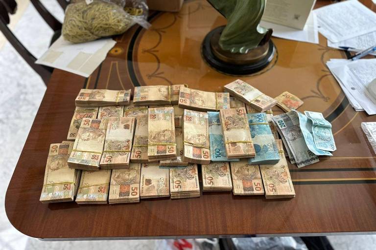 Diversas cédulas de real empilhadas em grupos sobre uma mesa de madeira com detalhes decorativos. Ao fundo, objetos como uma escultura verde, papéis e sacolas estão visíveis.
