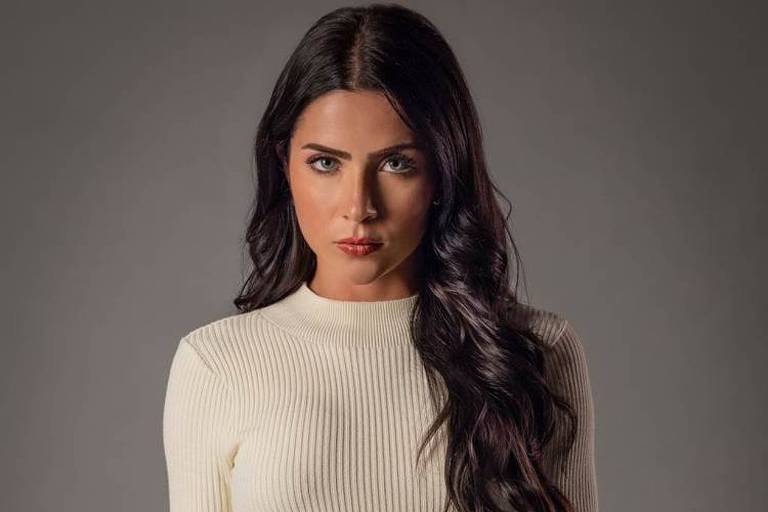 Mulher de cabelos longos e ondulados, escuros, veste blusa branca de malha canelada, posando contra fundo cinza neutro.