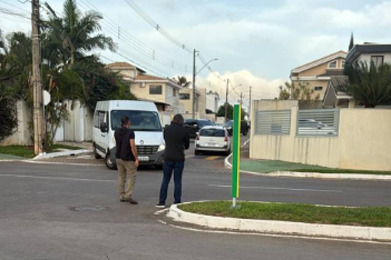 Duas pessoas estão paradas no meio de um cruzamento em bairro residencial. Uma delas segura uma câmera, possivelmente filmando ou fotografando. Ao fundo, há uma van branca estacionada e casas com muros baixos e vegetação, incluindo uma palmeira. O céu está parcialmente nublado.
