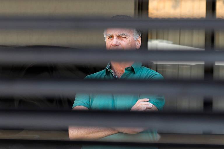 Homem de meia-idade com camisa verde de mangas curtas está com os braços cruzados, visto através de persianas horizontais que parcialmente obstruem a visão.