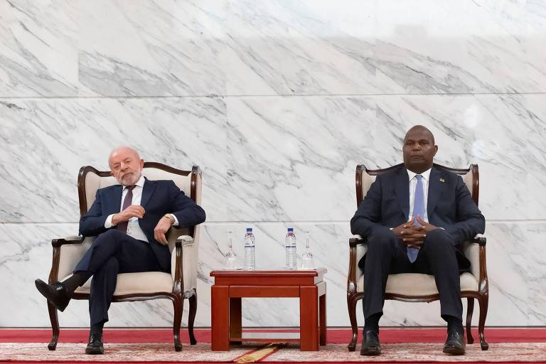 Dois homens sentados em cadeiras de madeira com estofado claro, separados por uma mesa pequena com duas garrafas de água, em um ambiente com parede de mármore branco. À esquerda, a bandeira do Brasil; à direita, a bandeira de Moçambique. Acima deles, um brasão dourado fixado na parede.
