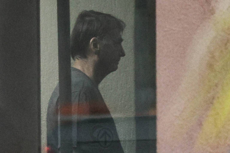 Homem de perfil, com cabelo curto e escuro, está em ambiente interno com iluminação baixa. Ele veste camiseta escura e está próximo a uma parede clara, com reflexos e sombras visíveis na imagem.