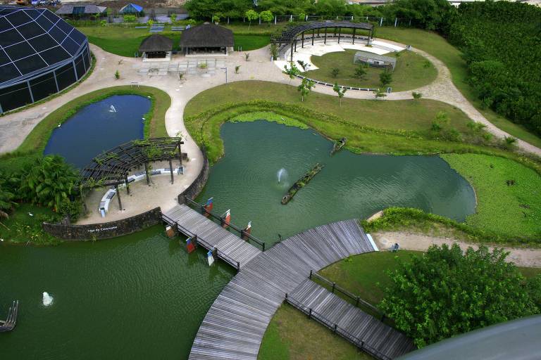 Vista aérea de parque com três lagoas conectadas por passarelas de madeira. Áreas gramadas cercam as lagoas, com bancos, pergolados e duas construções de palha. Caminhos de terra e vegetação densa delimitam o espaço.