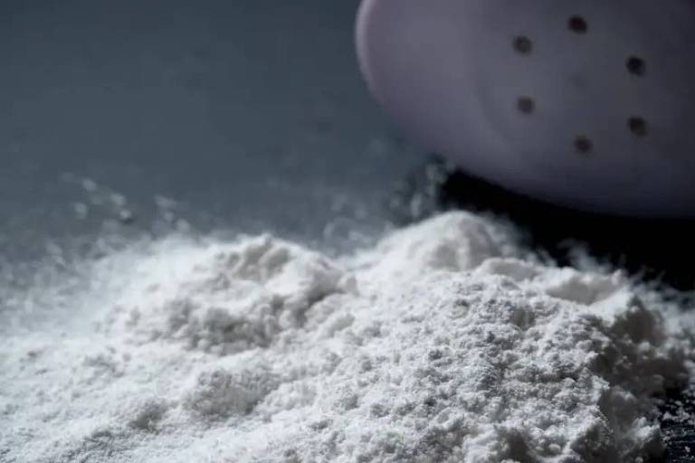 Pilha de farinha branca espalhada sobre superfície escura com peneira parcialmente visível ao fundo, sugerindo processo de peneiração.