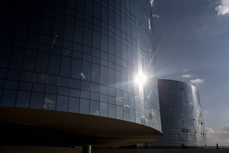 Dois prédios modernos com fachadas curvas de vidro refletem o céu azul e o sol forte. O sol aparece refletido no vidro do prédio à esquerda, criando um brilho intenso. O céu está parcialmente nublado com poucas nuvens.