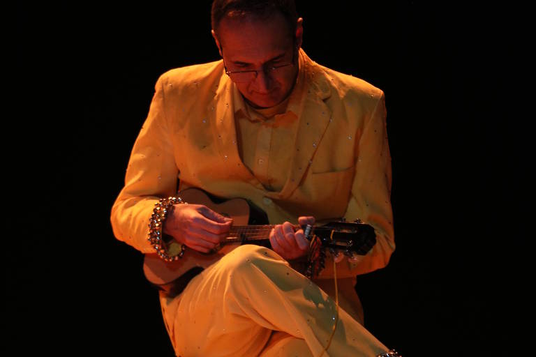Homem vestido com terno amarelo sentado em caixa, tocando instrumento de cordas pequeno, com fundo escuro e iluminação quente.