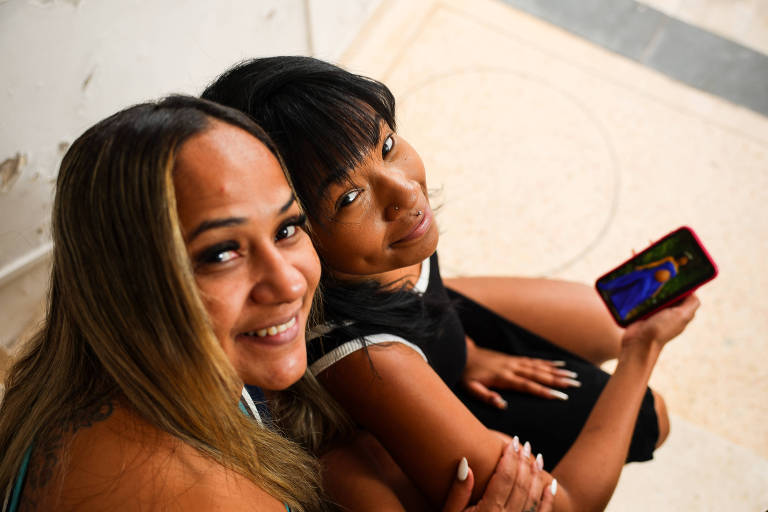 Duas mulheres sentadas lado a lado em escada interna, uma segura celular com tela colorida visível. Ambas sorriem e olham para a câmera.