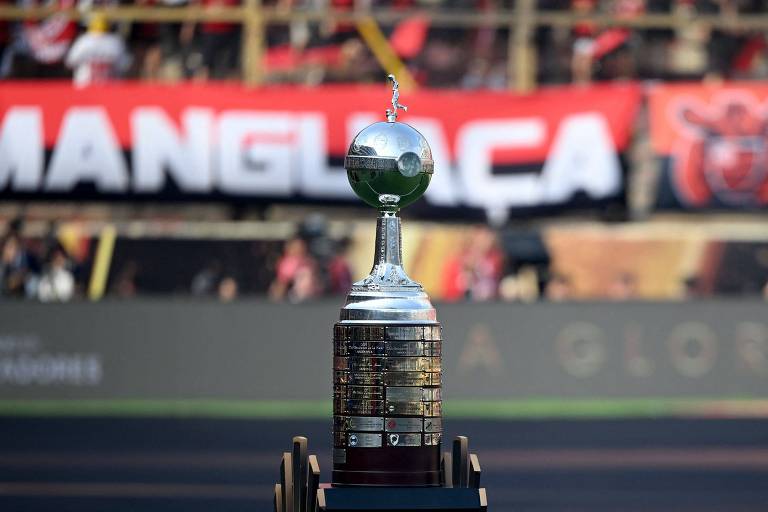 Troféu da Copa Libertadores em primeiro plano, com detalhes metálicos brilhantes e base escura, posicionado em campo de futebol. Ao fundo, torcida desfocada e faixa vermelha com letras brancas e pretas.