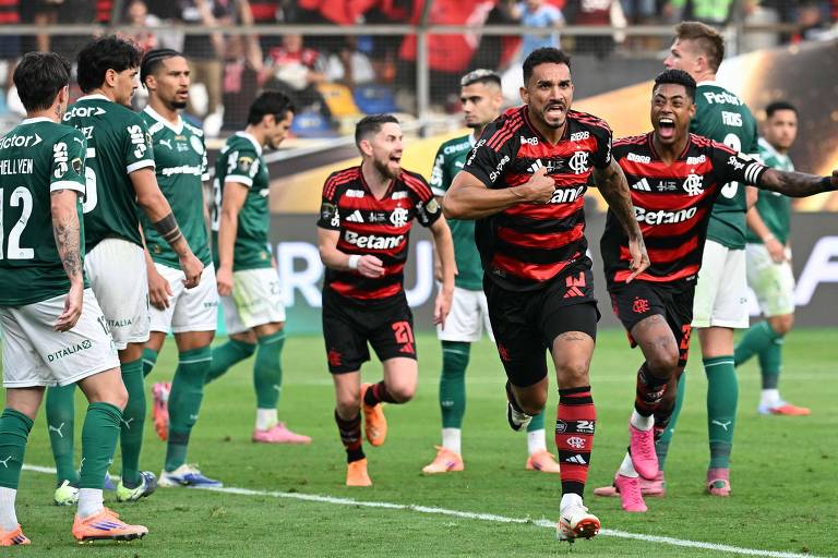 Jogadores do Flamengo vestindo uniforme vermelho e preto comemoram com expressão de alegria e braços abertos no campo. Jogadores do Palmeiras, com uniforme verde, observam a cena com semblante sério. O jogo ocorre em estádio com torcida ao fundo.