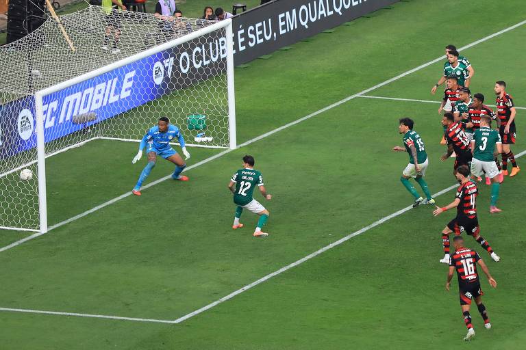 Goleiro em uniforme azul se prepara para defender bola durante cobrança de falta próxima à grande área. Jogadores de dois times, um com uniforme verde e outro com listras vermelhas e pretas, observam a jogada dentro da área.