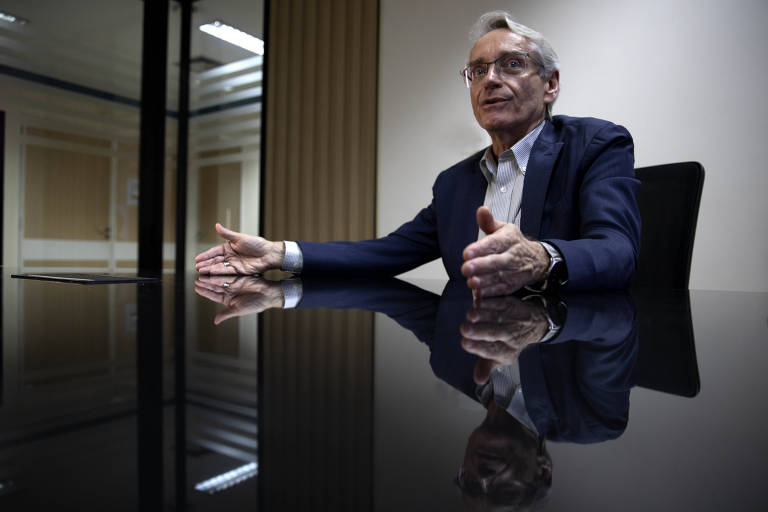 Homem idoso de cabelos grisalhos e óculos sentado à mesa de escritório escura, gesticulando com as mãos durante conversa. Ambiente interno com parede clara e janela com persianas ao fundo.