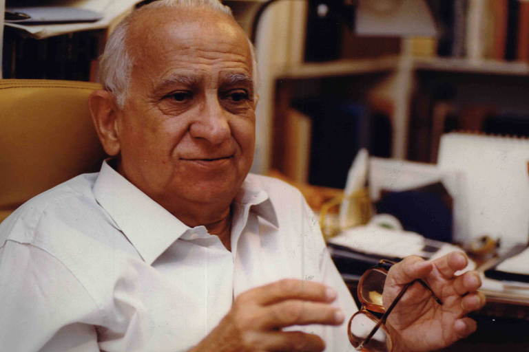 Homem idoso de cabelos grisalhos e camisa branca sentado em cadeira marrom em escritório. Ele segura um par de óculos com ambas as mãos, com estantes e papéis desfocados ao fundo.