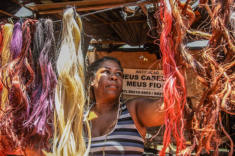 Mulher negra com blusa listrada observa fios coloridos pendurados em barraca de mercado. Fios em tons de vermelho, amarelo, roxo e marrom cercam a cena. Placa ao fundo indica nome da instituição e contatos.