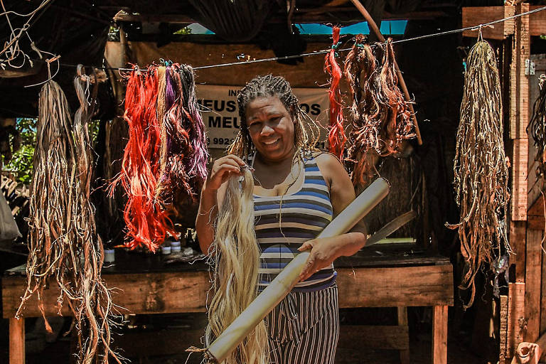 Mulher sorridente segura mechas longas de cabelo sintético e um tubo branco em barraca rústica. Ao fundo, várias extensões coloridas penduradas em cordas.