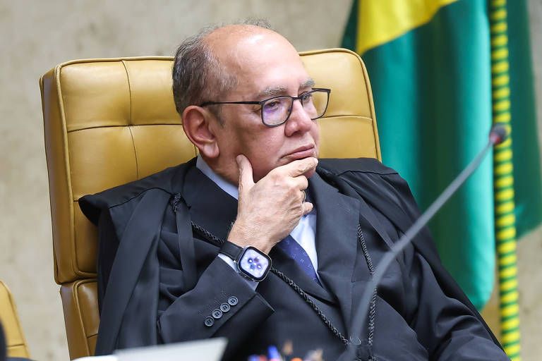 Ministro do Supremo Tribunal Federal sentado em cadeira de couro marrom, vestindo toga preta e óculos, com mão no queixo em gesto pensativo. Bandeira do Brasil ao fundo.