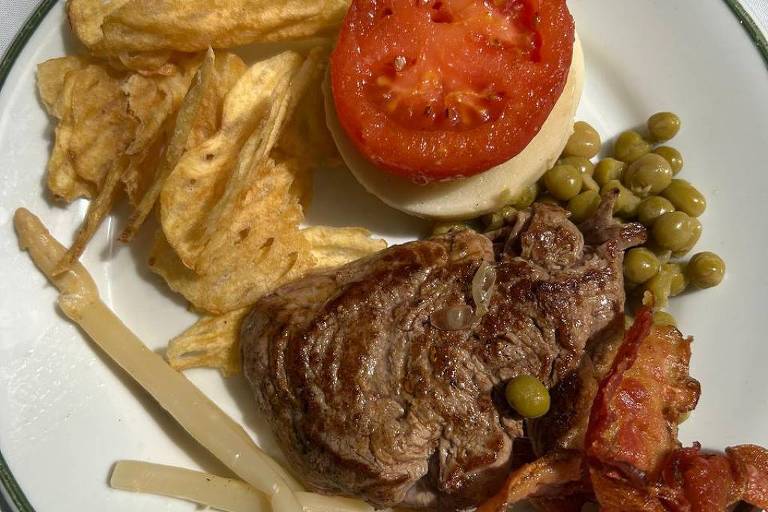 A white plate with green borders contains a piece of roast beef, slices of bacon, peas, a slice of tomato on a cheese slice and potato chips on the side.