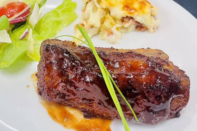 Costela ao molho escuro servida com molho no prato branco, acompanhada de salada verde com tomate e porção de gratinado de batata ao fundo.