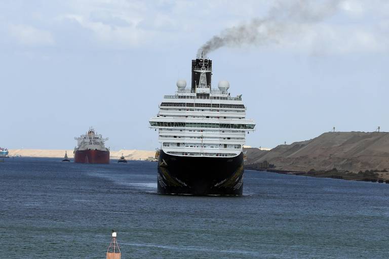Navio de cruzeiro grande navegando em canal estreito com fumaça saindo da chaminé. Ao fundo, navio cargueiro vermelho se aproxima. Margens do canal com terreno arenoso e céu parcialmente nublado.