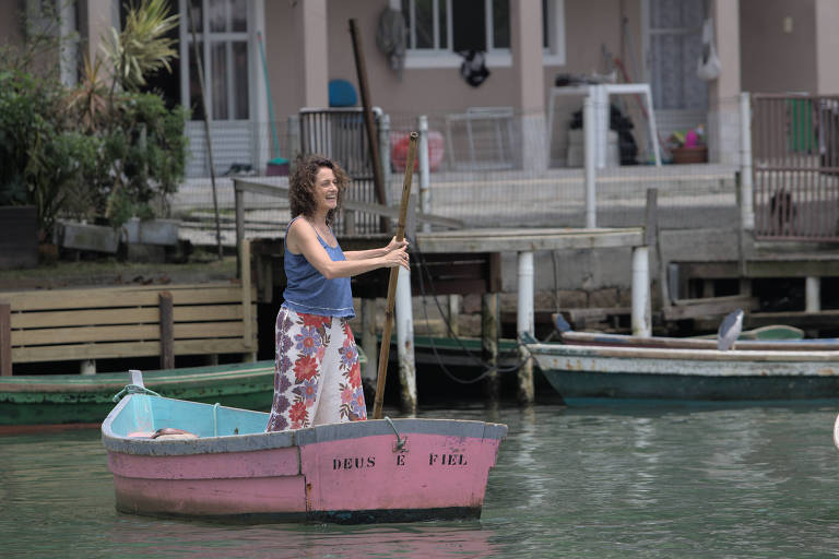 Woman with curly hair, dressed in a blue blouse and floral skirt, rows a pink boat with the inscription 