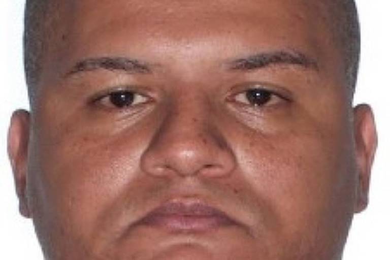 Homem de pele morena clara, cabeça raspada, sem cabelo visível, com expressão facial neutra, olhando diretamente para a câmera. Fundo branco neutro.