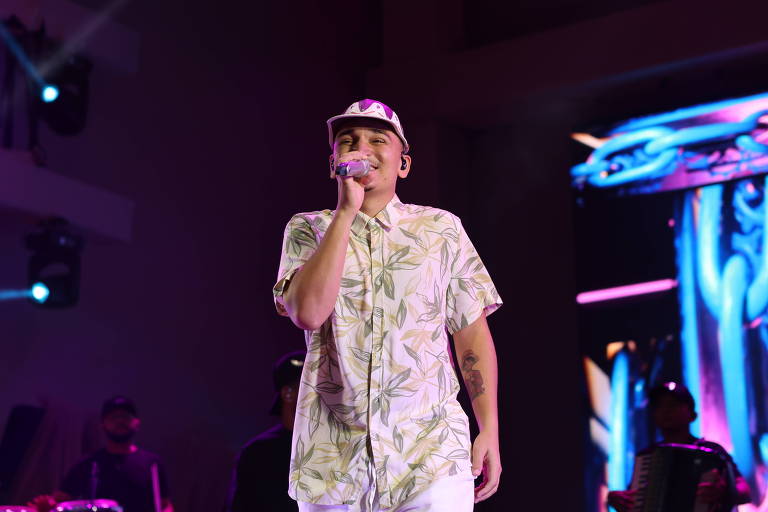 Cantor veste camisa clara com estampa de folhas e boné, segurando microfone nary centro bash palco. Ao fundo, músicos tocam instrumentos e há iluminação colorida roxa e azul, com tela integer exibindo imagens abstratas.