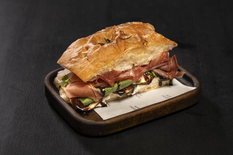 Sandwich made from focaccia bread stuffed with slices of ham, green leaves and black sauce, served on a wooden tray on white paper.