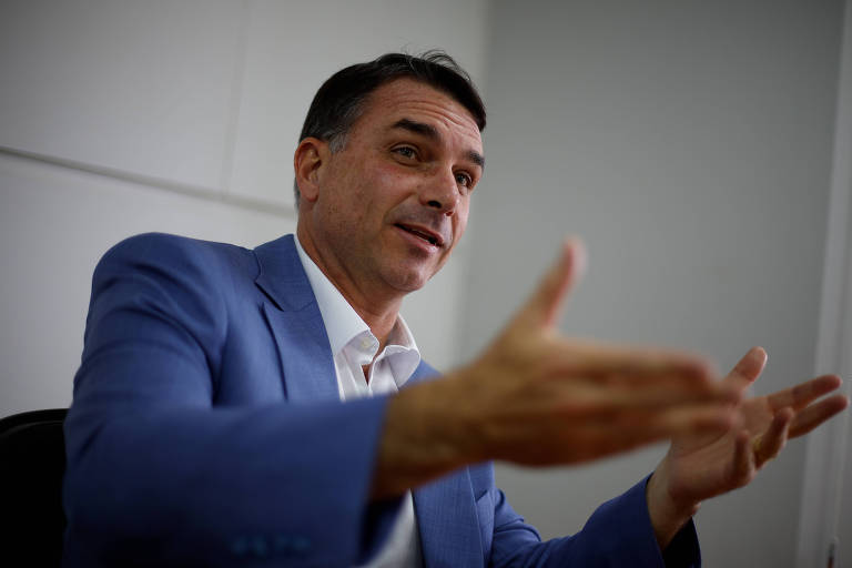 Homem de meia-idade veste terno azul e camisa branca, gesticulando com as mãos abertas em ambiente interno com parede branca ao fundo.