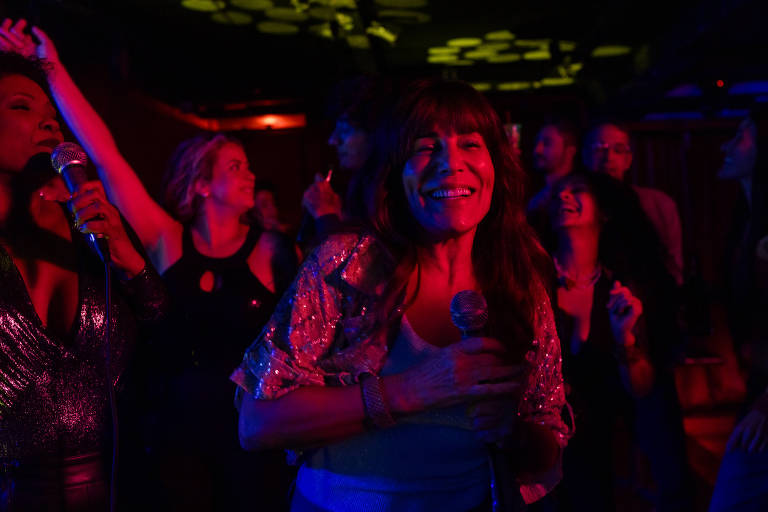Group of people in a dark environment with colorful lighting, some holding microphones and others dancing. A woman in the center smiles while holding a microphone, wearing a light blouse. Background with more moving people and decorative lights on the ceiling.