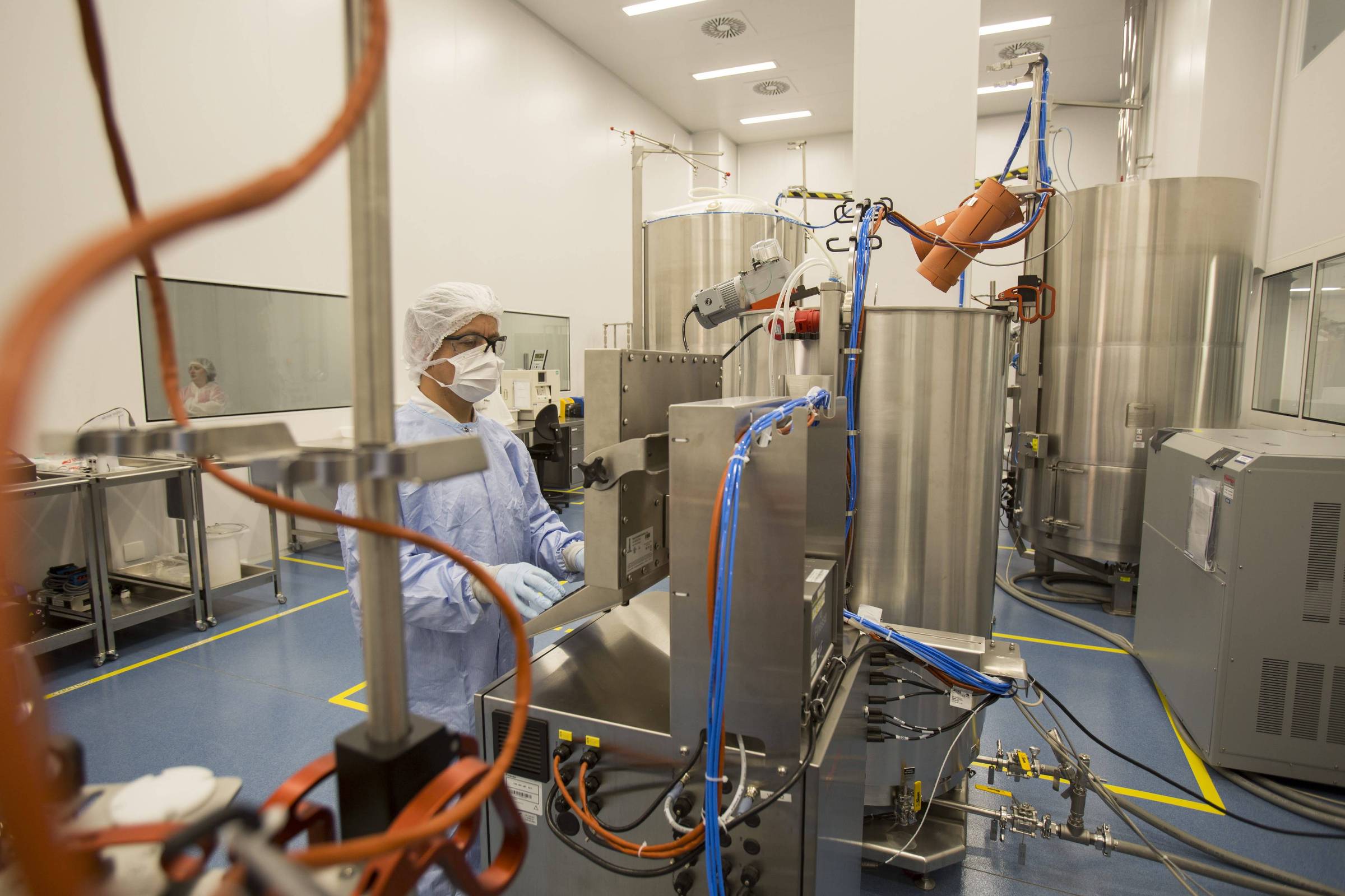 Profissional com roupa de proteção completa, máscara e touca, manuseia máquina em ambiente limpo com tanques metálicos e tubulações. Piso azul e paredes brancas indicam área controlada.