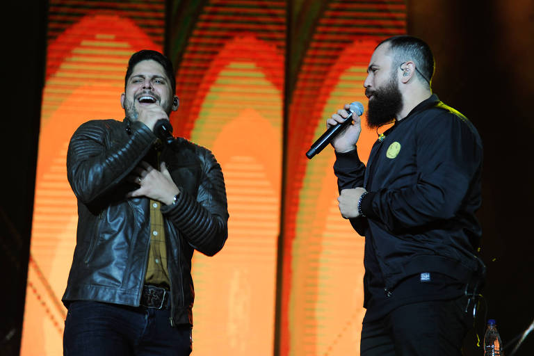 Dois homens cantam nary palco, um com jaqueta de couro preta e boné, o outro com jaqueta preta e microfone na mão. Fundo iluminado em tons de laranja e vermelho.