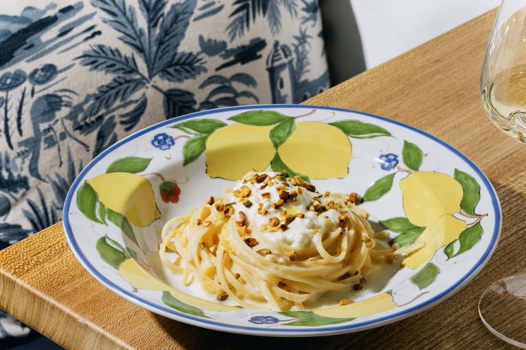 Porção de espaguete ao molho branco servida em prato decorado com limões e folhas verdes. Massa coberta por creme branco e nozes picadas, sobre mesa de madeira com toalha estampada ao fundo.