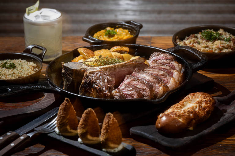 Sliced roast beef served in an iron skillet with potatoes on the side. Side dishes include rice, farofa, pumpkin puree and cheese bread. A clear drink with ice and lemon complements the wooden table.