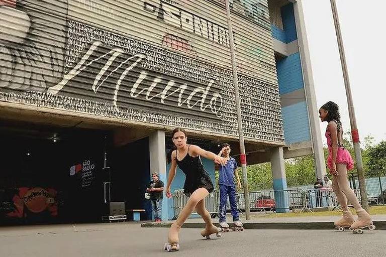 Três jovens patinadoras em patins de rodas praticam em frente a um prédio com fachada grafitada. Uma delas, no centro, usa vestido preto e está em movimento, enquanto as outras duas observam e se preparam para patinar. O ambiente é urbano, com carros estacionados e vegetação ao fundo.