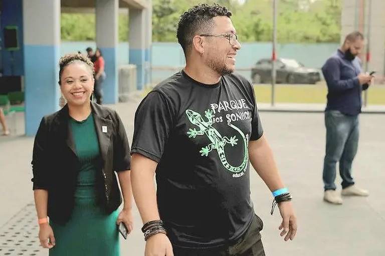 Homem de camiseta preta com estampa de lagarto e mulher de vestido verde com blazer preto caminham lado a lado em área externa coberta. Ao fundo, homem usa máscara e olha para celular.
