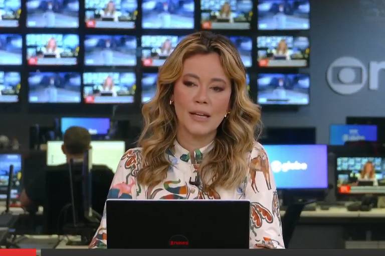 Apresentadora de telejornal sentada à frente de um laptop em estúdio com múltiplas telas ao fundo exibindo imagens diversas. Ambiente tecnológico com pessoas trabalhando em computadores ao fundo e logo da emissora 'GloboNews' visível na parede.