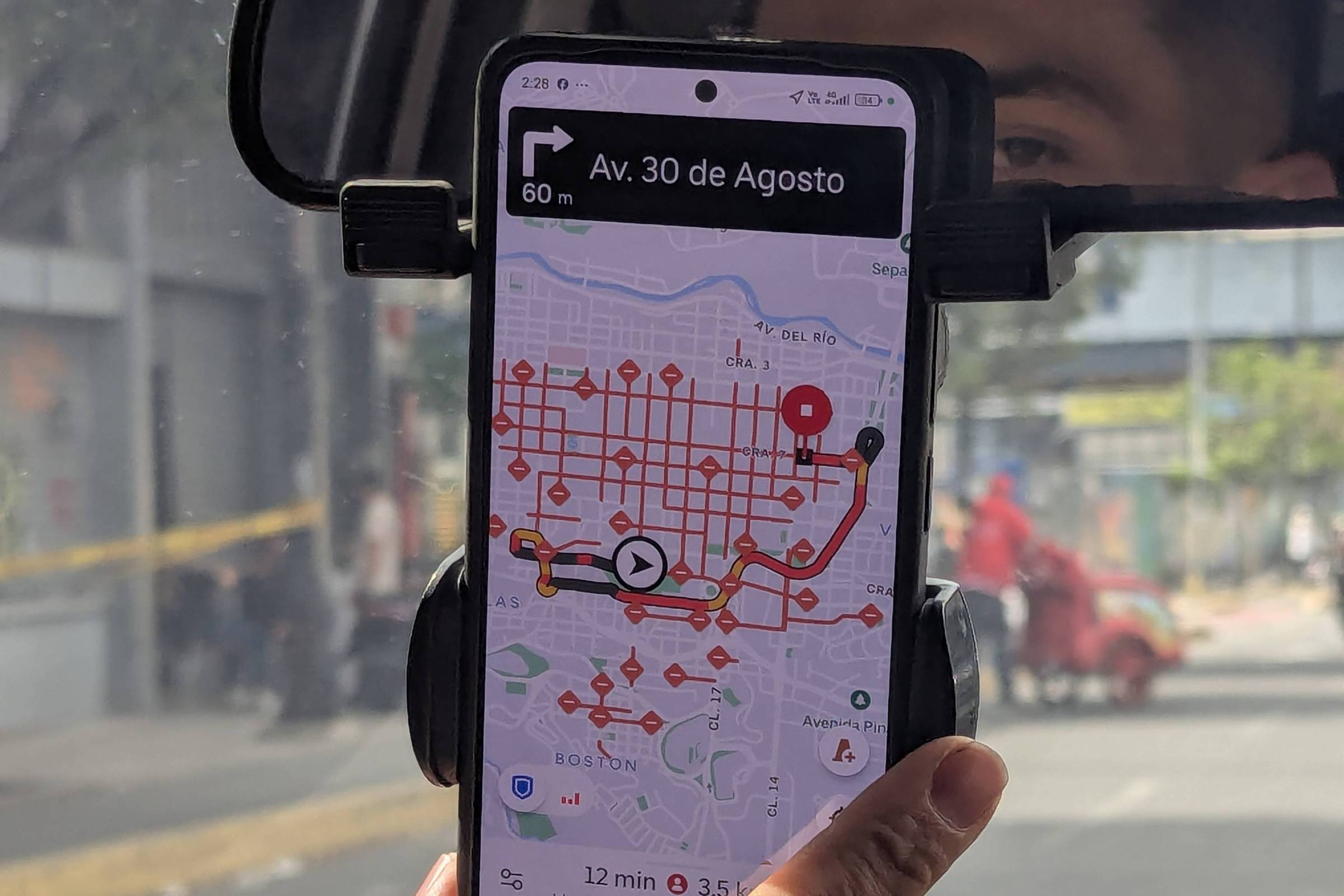 Mão segura smartphone com aplicativo de navegação aberto, mostrando rota em mapa com pontos vermelhos. Tela indica destino na Av. 30 de Agosto. Interior do carro e rua com pedestre ao fundo visíveis.