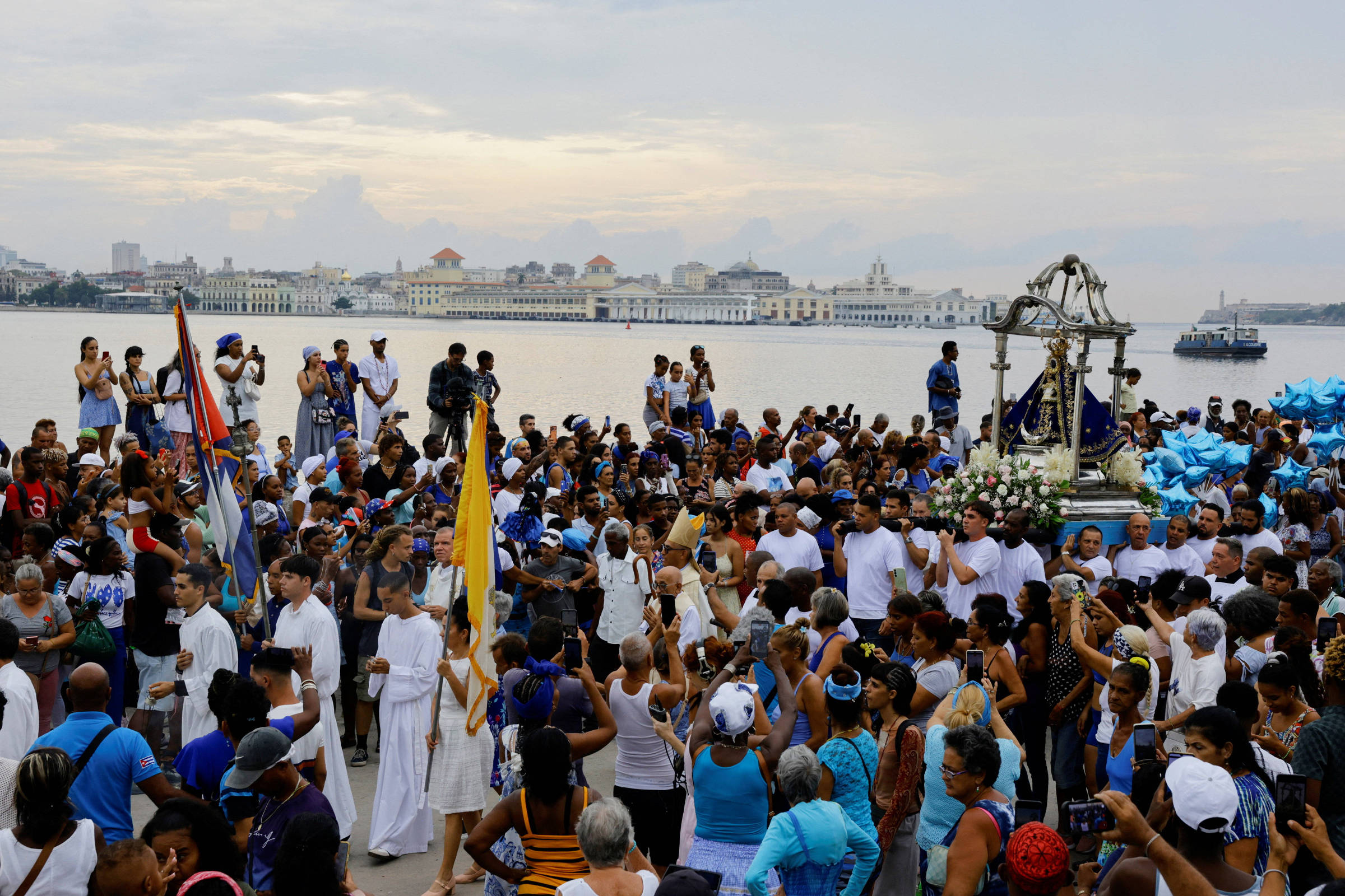 Centenas de pessoas participam de procissão religiosa à beira-mar, com imagem de santo em andor decorado com flores brancas. Muitos vestem branco e azul, e seguram balões azuis. Ao fundo, edifícios históricos e barco no mar sob céu nublado.